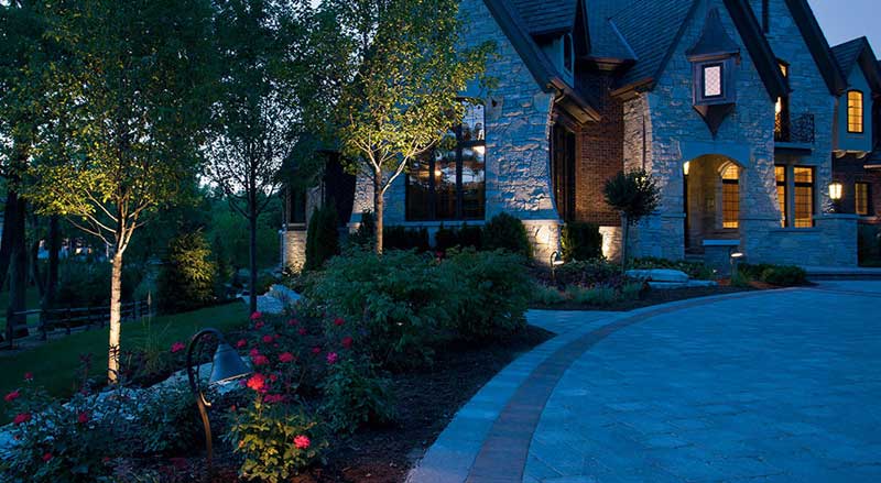 Warm landscape lighting illuminating trees and flower beds along a stone home’s driveway, creating dramatic nighttime curb appeal