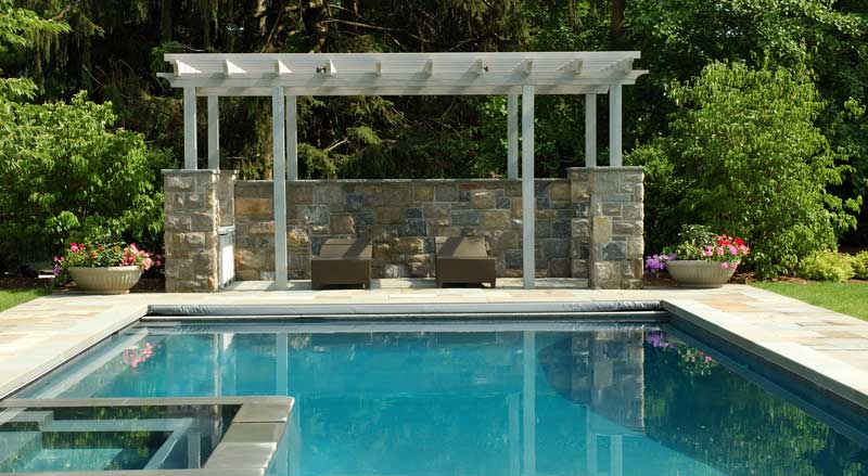 Modern pergola with stone backdrop and lounge chairs beside a serene backyard pool, showcasing luxury poolside landscape design in New Jersey.