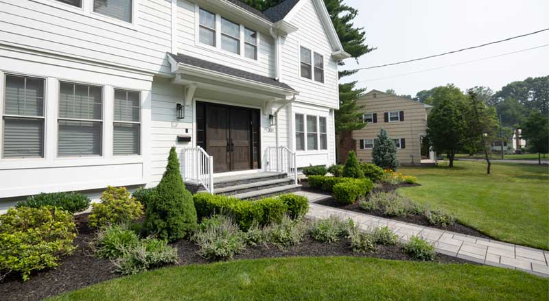Front yard landscaping with manicured shrubs, flower beds, and a stone walkway leading to a modern New Jersey home