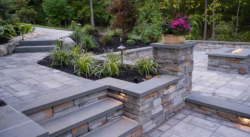Elegant stone steps and seating walls with accent lighting, surrounded by garden beds and a fire feature in a landscaped backyard
