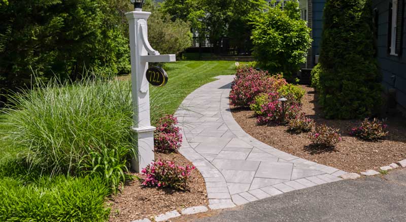 Curved stone walkway bordered by flowering shrubs, ornamental grasses, and mulch leading to the entrance of a New Jersey home