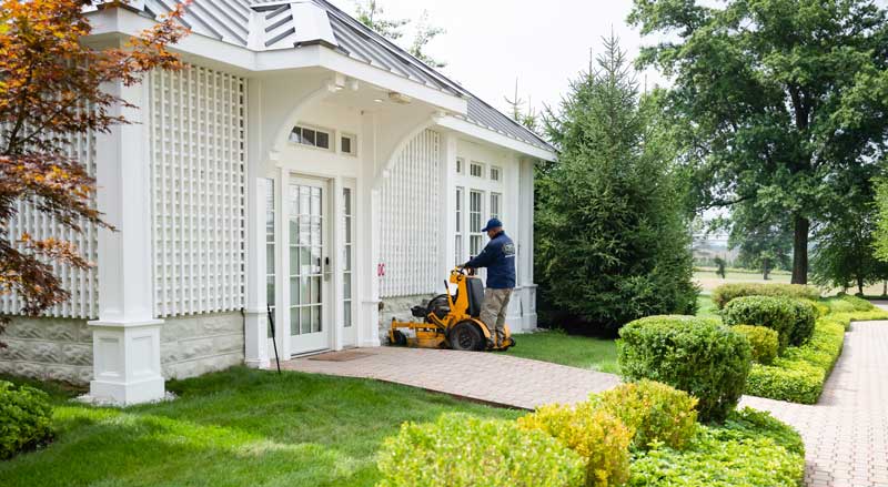 Professional landscaper using a ride-on mower to maintain grass near a clubhouse entrance in a multi-family HOA property