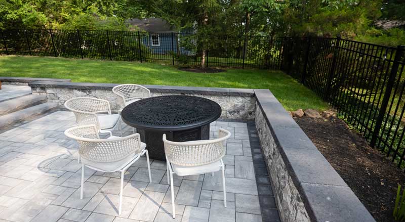 A paver patio with a firepit with four chairs and a seating wall