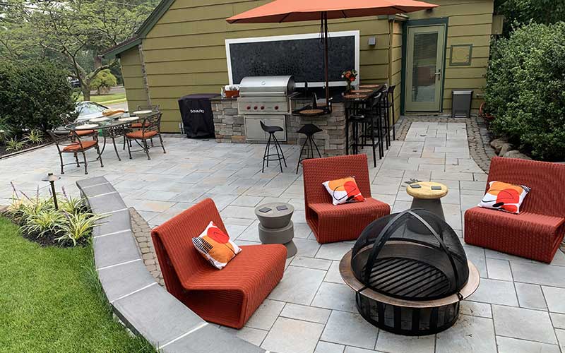 Paver patio with separate entertaining spaces for dining, comfortable seating around a firepit, a bar, and kitchen.