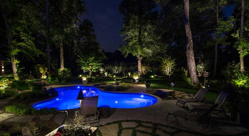 Glowing inground pool surrounded by lush, professionally lit landscaping,