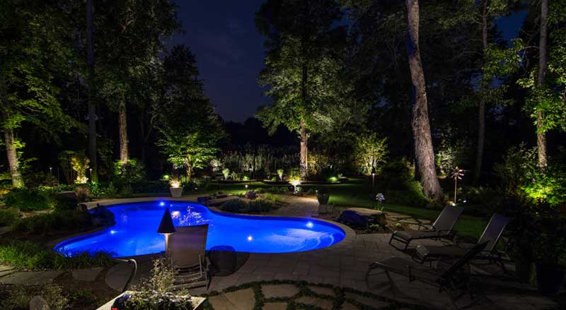 Backyard poolscape with landscape lighting, natural stone patio, and mature trees creating a luxury outdoor retreat for NJ summer evenings.