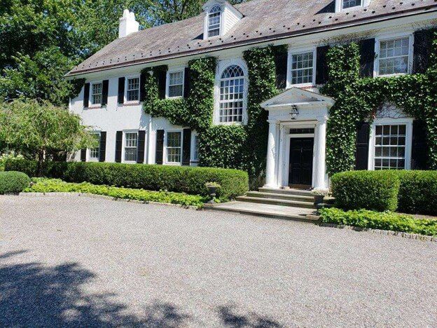 A prominent white house with contrasting black shutters, enveloped in ivy, highlighting the importance of continuous landscape maintenance