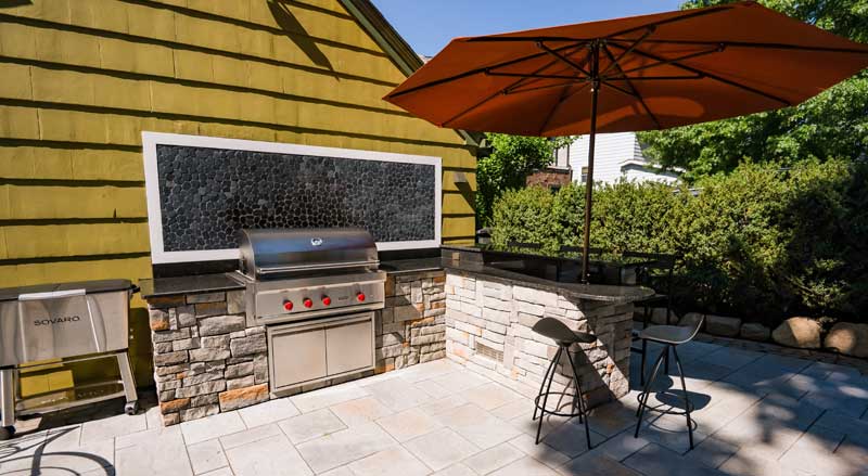 Custom stone outdoor kitchen with built-in stainless steel grill and bar seating, illustrating functional cooking spaces as a NJ landscaping trend.