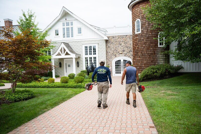 Two landscape workers walk along a path towards a large residence that has a well-maintained property, the property itself highlights the importance and value of professional landscape maintenance