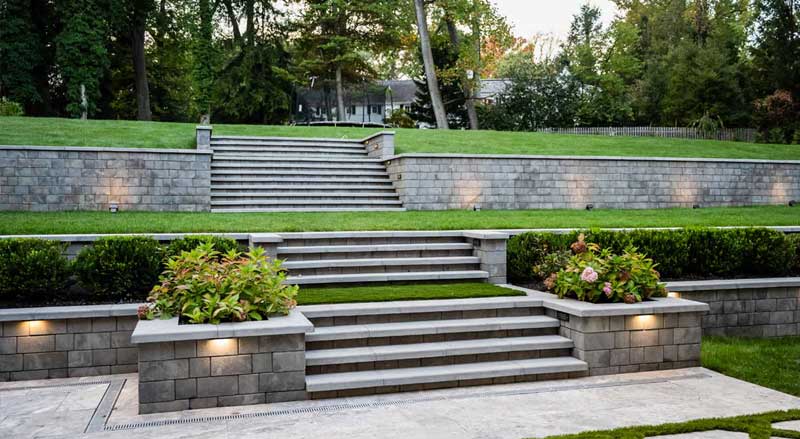 Tiered stone retaining walls and steps with landscape lighting and manicured lawn in a professionally designed backyard